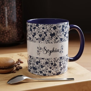 Puede incluir: Una taza de cerámica blanca con un estampado floral azul y un asa azul. La taza tiene el nombre "Sophia" impreso. Una cuchara y un pastel están sobre una superficie de madera.