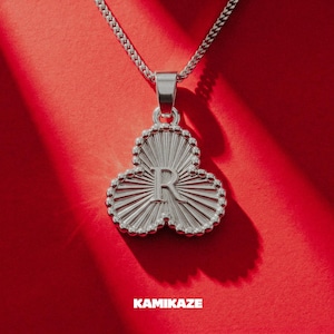 Puede incluir: Collar con colgante plateado con un diseño de trébol de tres hojas, con la letra "R" en el centro. El collar cuelga sobre un fondo rojo vibrante. La palabra "KAMIKAZE" se muestra en la parte inferior.