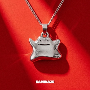 Puede incluir: Collar con colgante plateado con el diseño del personaje Ditto. El colgante, con forma de mancha sonriente, cuelga de una cadena plateada. El fondo es rojo liso, con la palabra "KAMIKAZE" en blanco en la parte inferior.