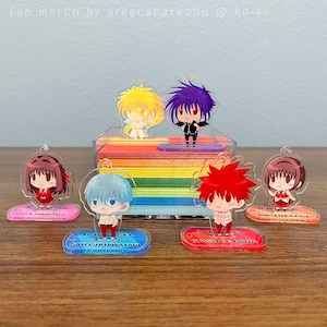 May include: Acrylic anime character charms with colorful bases. The charms feature various characters with different hair colors and outfits. The bases have character names and the text "D.N. Angel". The background includes a rainbow-colored block.