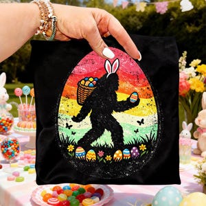 May include: Black tote bag featuring a Bigfoot silhouette wearing bunny ears and carrying a basket of eggs. The design is set against a colorful rainbow egg shape with Easter eggs and flowers at the bottom.