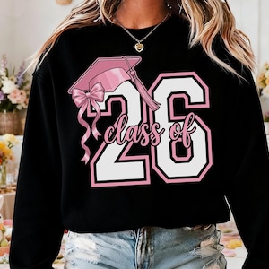 May include: Black sweatshirt featuring a pink graduation cap graphic and the text "Class of 26" in white with a pink outline. The design includes a pink bow and tassel. A casual garment.