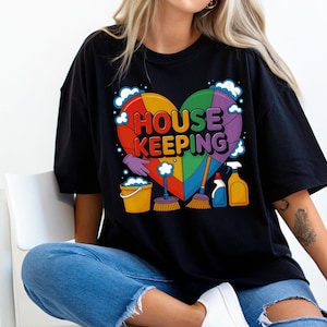 Puede incluir: Camiseta negra con un diseño de corazón arcoíris que dice "HOUSE KEEPING". El diseño del corazón incluye utensilios de limpieza como un cubo, escobas y pulverizadores. La camiseta tiene un ajuste holgado.