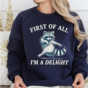 Könnte beinhalten: Marineblaues Sweatshirt mit einer Waschbär-Illustration und dem Text "FIRST OF ALL I'M A DELIGHT" in Weiß. Der Waschbär wird in einer Tanzpose dargestellt. Das Sweatshirt ist einfarbig und hat einen Rundhalsausschnitt.