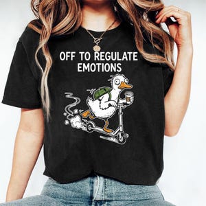 May include: Black t-shirt with the text "OFF TO REGULATE EMOTIONS." The shirt features a cartoon duck riding a scooter, carrying a backpack and a coffee cup. The duck is white with an orange beak.