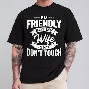 May include: Black t-shirt with white text: "--- I'M --- FRIENDLY BUT MY Wife ISN'T DON'T TOUCH". The text is in a bold, clear font. The shirt is a classic crew neck style, suitable for casual wear.
