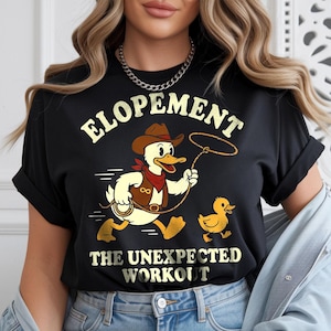 May include: Black t-shirt with a cartoon duck wearing a cowboy hat and vest, holding a lasso, and running with a smaller duckling. The text "ELOPEMENT" and "THE UNEXPECTED WORKOUT" are printed in gold.