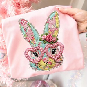 以下が含まれることがあります： ハート型のメガネをかけたウサギの顔が刺繍されたピンク色の布製品。ウサギは花柄の耳と花の王冠を持っています。メガネは真珠で縁取られています。ウサギの顔は黄色、青、ピンクです。