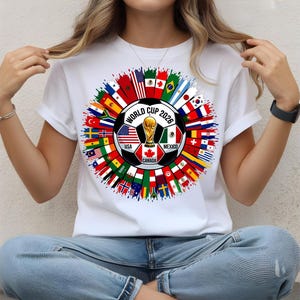 Op de afbeelding: Wit T-shirt met een kleurrijk grafisch ontwerp voor het Wereldkampioenschap 2026. Het ontwerp toont een voetbal met vlaggen van deelnemende landen, de Wereldbeker-trofee en de tekst "WORLD CUP 2026".