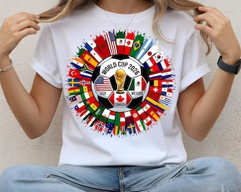 May include: White t-shirt featuring a colorful graphic design for the 2026 World Cup. The design includes a soccer ball with flags of participating countries, the World Cup trophy, and the text "WORLD CUP 2026".