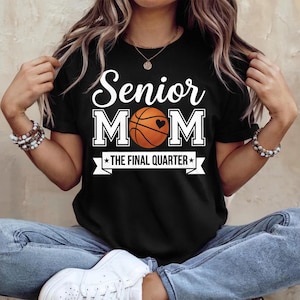 May include: Black t-shirt with the text "Senior Mom" in white block letters above a basketball graphic with a heart. Below the basketball is a banner that reads "THE FINAL QUARTER" in white.