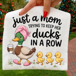 Op de afbeelding: Wit grafisch T-shirt met een cartoon eend met een roze handdoek en slippers, met een koffiekopje. Tekst luidt: "just a mom trying to keep her ducks in a row." Drie gele eendjes volgen.