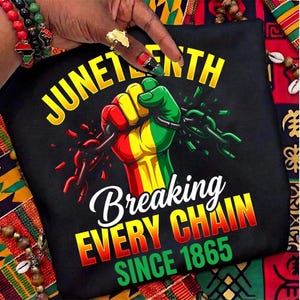 May include: Black t-shirt featuring a fist breaking chains, coloured in red, yellow, and green. The words "Juneteenth" and "Breaking Every Chain Since 1865" are in yellow, white, and green.