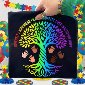 Può includere: T-shirt nera con un design ad albero colorato e le parole "Diversità, Gentilezza, Pace, Qualità, Amore, Inclusione, Speranza". L'albero è nei colori dell'arcobaleno, con impronte di mani e pezzi di puzzle. Palloncini e decorazioni a forma di puzzle sullo sfondo.