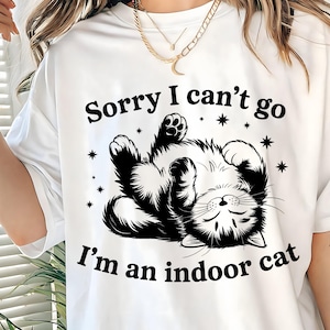 以下が含まれることがあります： 「Sorry I can't go, I'm an indoor cat」の文字と、仰向けに寝ている猫の白黒イラストがプリントされた白いTシャツ。星のマークが周りに描かれています。