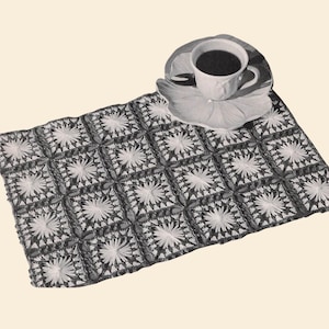 May include: A crocheted placemat with a floral design, in shades of gray and white, is shown with a cup of coffee on a matching saucer. The placemat is made of square motifs. The image includes the text "Knot Forgotten Patterns".
