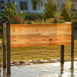 May include: A raised wooden planter box with black metal legs. The planter is filled with a variety of flowers and plants. The planter is made of natural wood and has a light brown finish. The planter is sitting on a patio with a stone floor.