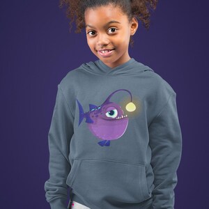 May include: A hooded sweatshirt in a muted blue-gray color featuring a cartoon anglerfish design. The anglerfish is purple with a glowing yellow lure. The sweatshirt has a front pocket and is worn by a person.