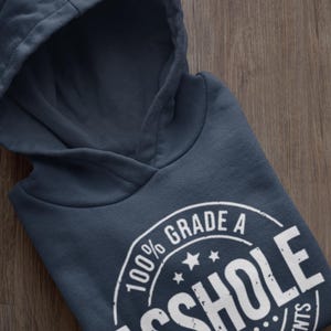 May include: Navy blue hoodie with a hood, folded on a wooden surface. The hoodie features a white graphic that reads "100% Grade A Asshole" in a circular design with stars and text. The hoodie is made of a soft material.