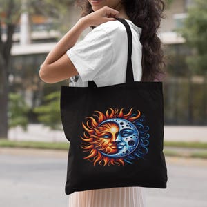 May include: A black tote bag featuring a vibrant graphic print of a sun and moon face. The sun side displays warm orange, yellow, and red flames, while the moon side features cool blue tones with craters and stars, creating a celestial design.