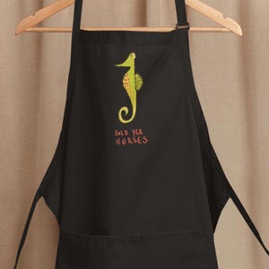 May include: A black apron featuring a graphic of a yellow seahorse with orange spots and the text "HOLD YER HORSES" printed in orange below it. The apron includes a neck strap, two long side ties, and a large front pocket for storage.