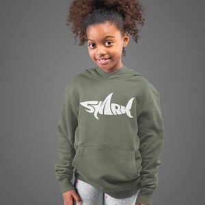 May include: A young person wearing an olive green hoodie with a white shark design. The hoodie has a front pocket and a hood. The person is also wearing gray leggings with a pink heart pattern.