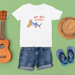 May include: A white t-shirt with the text "HOT DOG SUMMER" and a cartoon shark eating a hot dog. The image also includes denim shorts, a straw hat, blue flip-flops, and a ukulele on a green background.