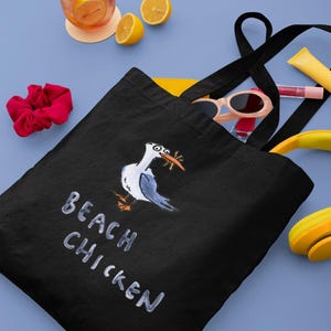 May include: Black tote bag with a cartoon seagull illustration and the text "BEACH CHICKEN" in white. The bag is on a blue surface with a drink, lemons, sunglasses, and other accessories.