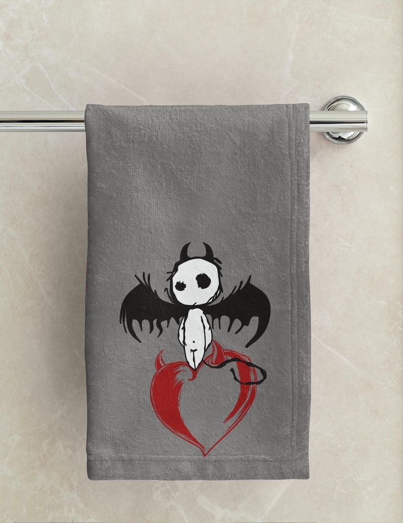 May include: Gray bath towel with a cartoon devil design. The devil has black wings, a white body, and a red heart. The towel is hanging on a chrome towel bar.