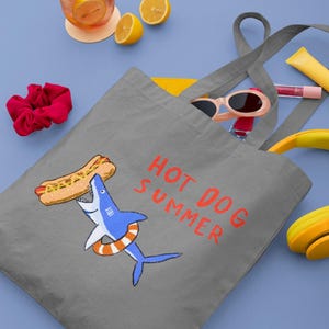 May include: A gray tote bag with a shark graphic holding a hot dog, with the text "HOT DOG SUMMER" in red. The bag is filled with sunglasses, lip gloss, and a banana. A drink with lemon slices and a red scrunchie are nearby.