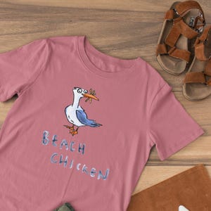 May include: A dusty rose t-shirt featuring a cartoon seagull with a bug in its beak. The text "BEACH CHICKEN" is below the bird in a light blue font. Brown leather sandals are next to the shirt.