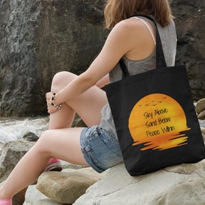 May include: A black tote bag featuring a graphic of a bright orange sunset over water with a flock of birds flying above. The text "Sky Above, Sand Below, Peace Within" is printed in black letters across the center of the circular sunset design.
