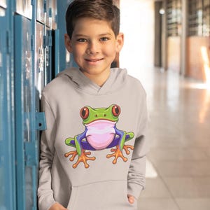 May include: A light gray hoodie featuring a colorful cartoon frog design. The frog has a green head, red eyes, a pink belly, and blue and purple limbs. The hoodie has a kangaroo pocket and is worn by a person.