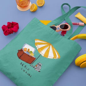 May include: A teal tote bag with a beach scene illustration. The illustration includes a cooler, a yellow and white striped umbrella, and a raccoon. The words "Beach Snacks" are printed on the bag. Sunglasses, lip gloss, and a scrunchie are visible.