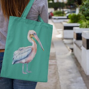 May include: A teal tote bag featuring a detailed illustration of a pelican with soft pink and white plumage. The bird is depicted in profile, facing right, with a long beak and light blue eye. The bag has two matching teal fabric shoulder straps.
