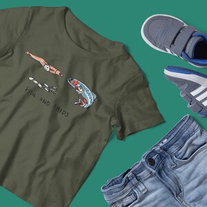 May include: Olive green t-shirt with a cartoon fish and chips design. The text "FISH AND CHIPS" is printed below the graphic. The image also includes a pair of gray sneakers and blue jeans.