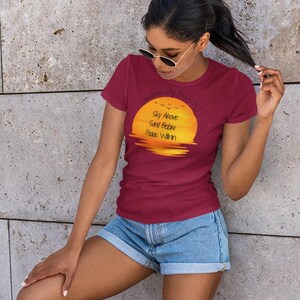 May include: A maroon t-shirt with the text "Dreams City Beach" and "Sky Above Sand Below Peace Within" printed on the front. The graphic is a yellow and orange sunset over water. The person is wearing blue jean shorts.