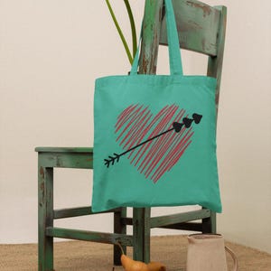 May include: A teal tote bag with a red heart pierced by a black arrow is displayed on a distressed green chair. The bag's design is a hand-drawn style. Pears and a pitcher are in the foreground.