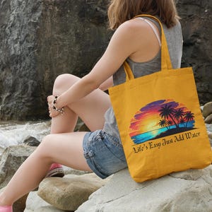 May include: A mustard yellow canvas tote bag featuring a colorful graphic of a sunset over the ocean with palm trees and the text "Life's Easy Just Add Water" printed in a black script font on the lower portion of the bag's front surface.