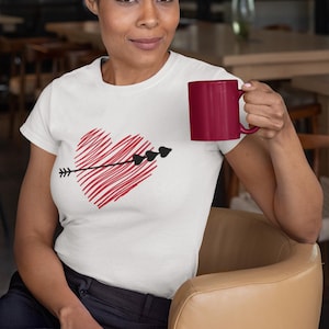 May include: A white t-shirt featuring a red heart pierced by a black arrow. The person in the image is holding a maroon mug. The shirt design is a simple, yet striking graphic. The overall theme is love and romance.
