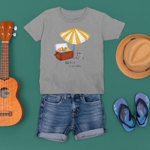 May include: A gray t-shirt with a beach scene illustration, denim shorts, a ukulele, a straw hat, and blue flip-flops. The shirt features a cooler, umbrella, and text that reads "Beach Snacks".