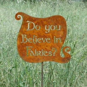 Puede incluir: Un letrero de jardín de metal oxidado naranja con la pregunta "Do you Believe in Fairies?" en letras verdes. El letrero está montado en un delgado poste de metal, colocado en una zona de césped, con dos grandes piedras en primer plano.