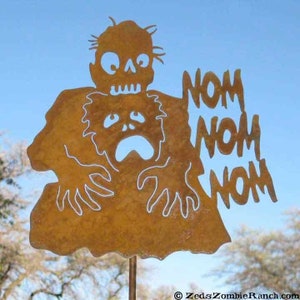 May include: A rusty metal zombie cutout with a smaller zombie inside its mouth. The zombie is holding the smaller zombie with its hands. The cutout has the word "NOM" written three times on the right side.