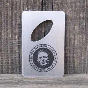 Peut inclure: Un ouvre-bouteille en argent avec un logo noir et blanc qui dit "Zeds Zombie Ranch.com Bisbee, Ariz."