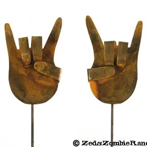 May include: Two rusty metal hand sculptures in the shape of the rock and roll hand gesture. Each hand is mounted on a thin metal rod.