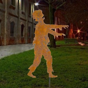 May include: A rusty metal silhouette of a zombie walking with its arms outstretched. The silhouette is standing on a grassy lawn in front of a brick building.