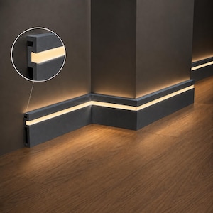 Puede incluir: Un rodapié gris oscuro instalado a lo largo de una esquina de pared, con un canal horizontal empotrado con iluminación LED blanca cálida integrada que proyecta un suave resplandor sobre el suelo de madera bajo el perfil.
