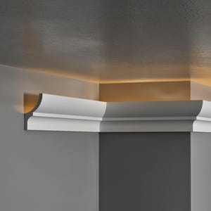May include: White crown moulding installed at the corner of a room. The moulding has a decorative profile and is positioned where the wall meets the ceiling. The walls are painted in shades of grey, and the ceiling has a warm, golden glow.