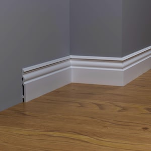 Puede incluir: Una esquina de una habitación con rodapiés blancos contra paredes grises y un suelo de madera. Los rodapiés tienen un perfil decorativo y están instalados donde la pared se encuentra con el suelo. El suelo tiene un veteado de madera cálido y natural.