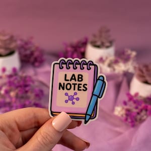 Może przedstawiać: Naklejka przedstawiająca różowy notatnik z czarnym napisem "LAB NOTES". Obok notatnika znajduje się niebieski długopis. Poniżej tekstu znajduje się fioletowa grafika cząsteczki. Tło obejmuje małe rośliny doniczkowe i fioletowe kwiaty.